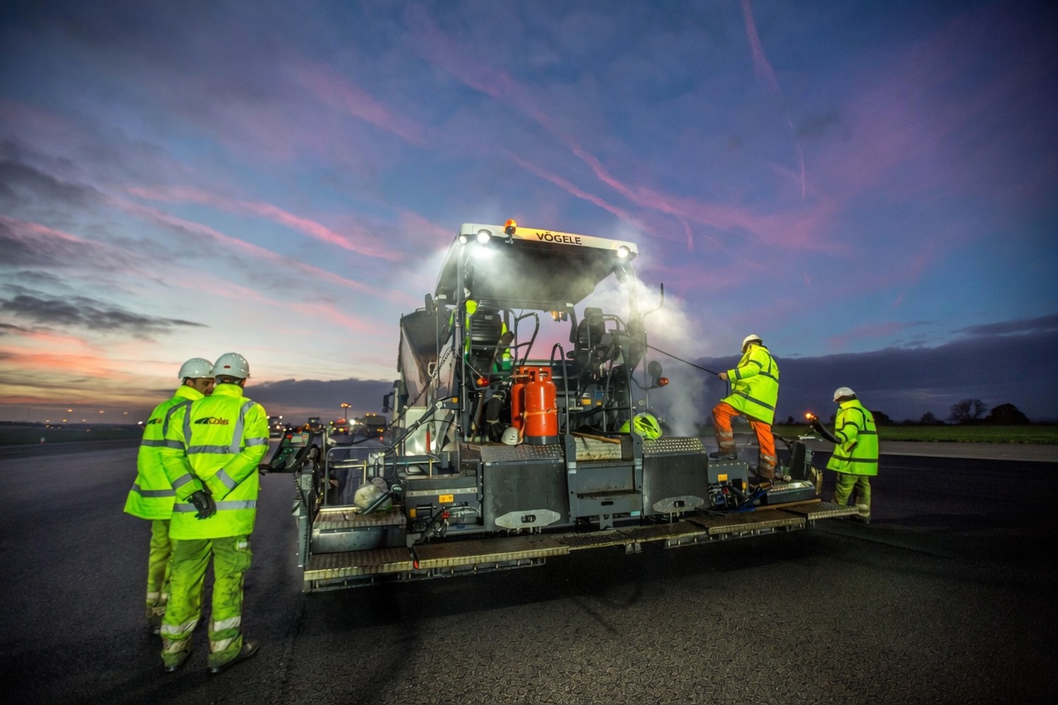 Night-shift surfacing crew working behind an asphalt paver under a dusk sky on a wide carriageway